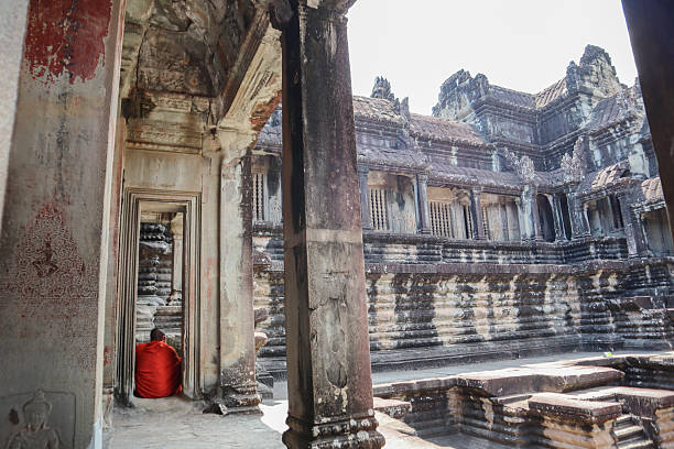 Siem Reap, Cambodia - April 18, 2025:A monk visits the UNESCO World Heritage Site of Angkor Wat. He holds a fan in his hand to cool off. Between the columns next to a building of the Angkor Wat temple complex, he stands still and looks at the impressive architecture.Angkor Wat is the most famous temple complex in the Angkor region of Cambodia. The temple is located about 240 km northwest of the capital Phnom Penh near Siem Reap, about 20 kilometers north of Lake Tonle Sap.In addition to the tourists, Buddhist monks are among the daily visitors to the temple.Angkor Wat functions as an outstanding national symbol that is representative of Khmer culture and the Cambodian people today. It can therefore be found as an illustration in a variety of state contexts, on the national flag, banknotes, etc. Even during the Khmer Rouge regime, a golden silhouette of the temple was part of the Cambodian flag.Angkor Temple is a majestic UNESCO World Heritage Site and a top destination for history enthusiasts traveling to Southeast Asia. Among the numerous temples in the Angkor complex, Angkor Wat stands out as the ultimate statement of Khmer architectural brilliance. It is the largest religious building in the world and is characterized by a perfect blend of symbolism and symmetry.