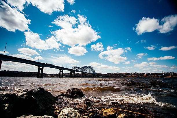 Photograph of the Julien Dubuque Bridge going over the Mississippi River in Dubuque, IA. Shot is a wide angle view with the shoreline in the foreground and a partly cloudy sky in the background.