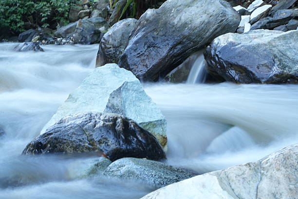 shallow stream flows over rocky bed, creating tranquil and natural scene. clear water, reflecting sunlight, moves around rocks, motion. brightness and blurred effect contribute to serene atmosphere