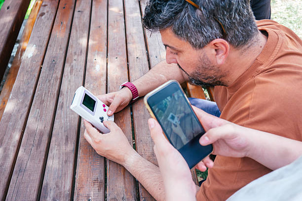A middle-aged man plays a nostalgic game on a retro console while a 14-year-old takes a photo or video with a smartphone, capturing a cross-generational tech moment.