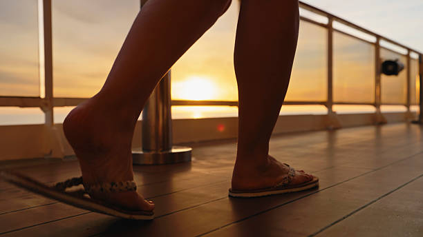 A female is leisurely walking on a cruise ship deck, wearing flip flops. The sun is setting, casting a warm glow over the scene. The atmosphere is serene and relaxed, perfect for a vacation setting.