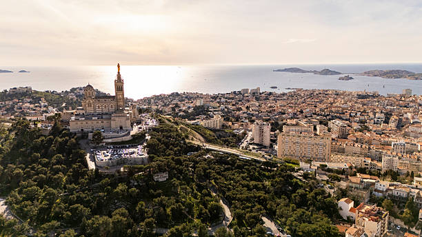 Notre-Dame de la Garde basilica is standing prominently on a hill overlooking the city of Marseille. The Mediterranean Sea is stretching in the background, while the urban landscape is sprawling below under a clear sky.