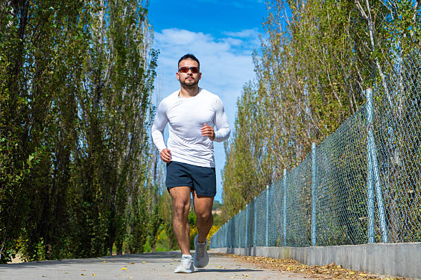 Hispanic man jogging outdoors on a sunny day wearing athletic clothes and sunglasses with trees and blue sky in the background for fitness concept.
