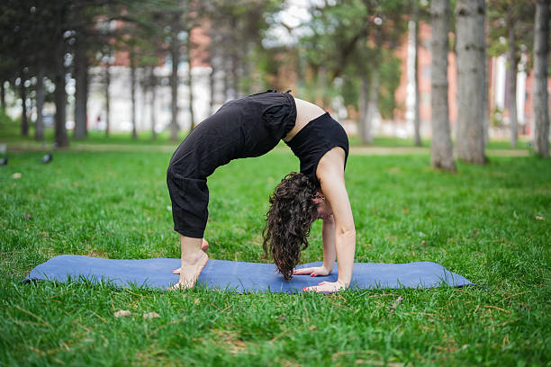 Young woman practicing advanced yoga pose in a park, performing upward bow (wheel) pose on a yoga mat surrounded by trees