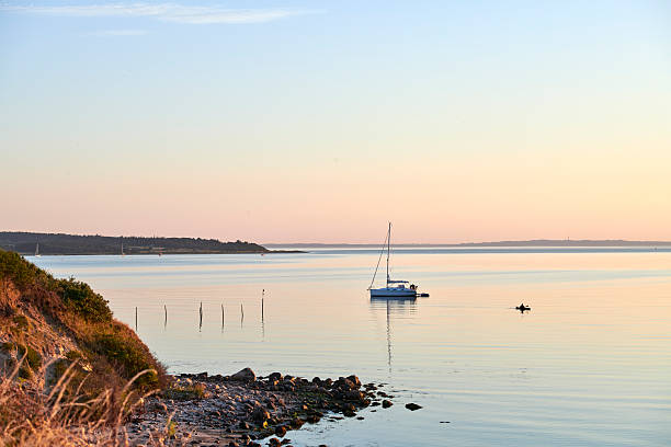 Experience the serenity of a sunset along the Danish coastline, featuring a sailboat and calm waters. The tranquil atmosphere beautifully showcases the scenic seaside, offering a moment of peace and reflection.