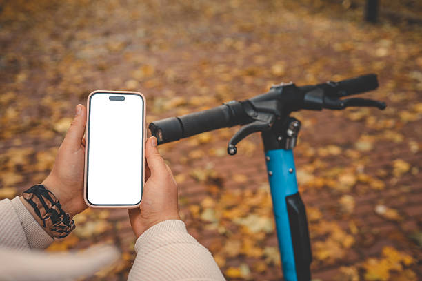 istock Person holding smartphone with blank screen next to electric scooter on a brick path covered with autumn leaves. Concept of navigation, mobility app, or ride-sharing in fall season 2214142220