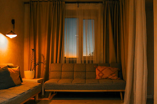 istock Cozy living room interior with warm lighting, grey sofas, decorative pillows, and a potted plant near the window during sunset 2214142261