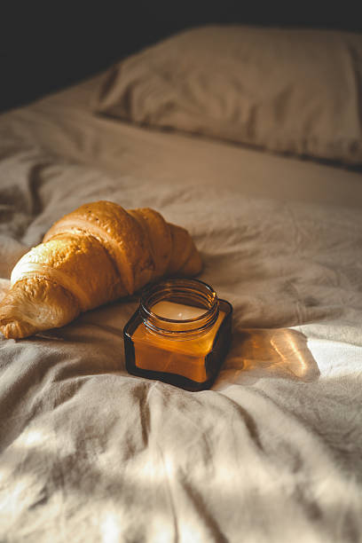 Fresh croissant and small amber glass candle jar placed on soft beige bedding, with golden morning sunlight casting warm shadows across the cozy bedroom setting
