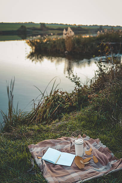 istock Open planner and reusable coffee cup on a plaid blanket near a peaceful lake at sunset, with fallen leaves adding an autumn touch 2214148314