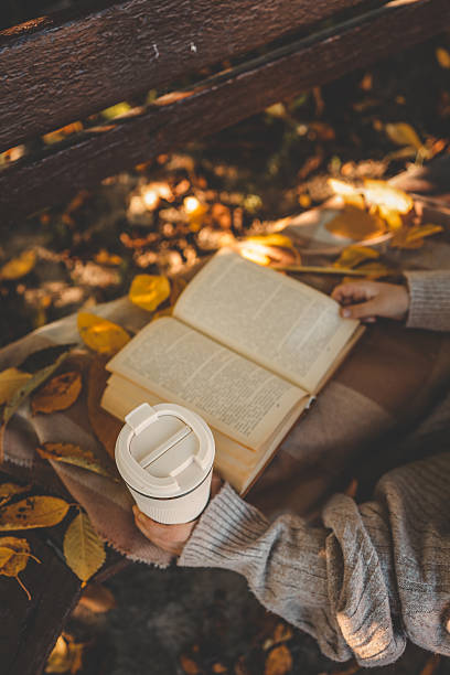 istock Person in cozy sweater holding reusable cup while reading a book outdoors in autumn park, surrounded by warm tones, fallen leaves, and soft sunlight 2214148324