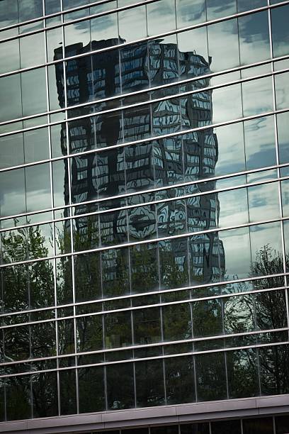 A modern glass building reflecting another structure, with trees in the foreground. The reflection creates an abstract pattern on the glass surface, showcasing urban architecture.