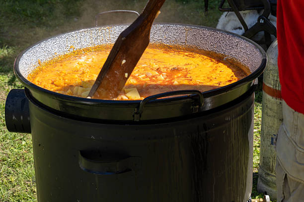 A large black cauldron filled with thick, bubbling stew is being cooked outdoors over an open flame. The hearty mixture of meat, beans, and vegetables creates a rustic, homemade atmosphere that evokes community gatherings and traditional outdoor cooking.