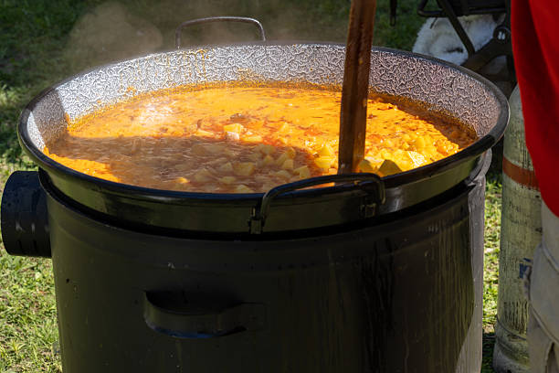 A person stirs a steaming stew of potatoes and meat in a large metal cauldron over an open gas flame during an outdoor event. The vivid yellow-orange color of the food and the rustic cooking method evoke a communal, festive atmosphere rooted in cultural traditions.