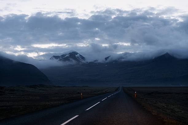 An empty asphalt road stretches into the distance, flanked by vast barren land and dramatic mountains partially veiled in mist and low-hanging clouds.