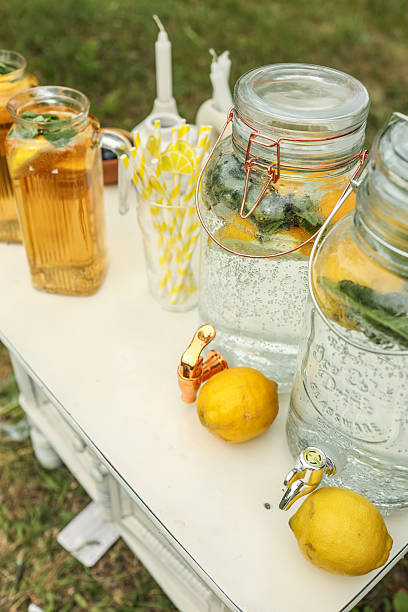 Glass beverage dispensers filled with lemon and mint-infused water, paired with striped paper straws and fresh lemons on a white outdoor table, perfect for a refreshing summer party station