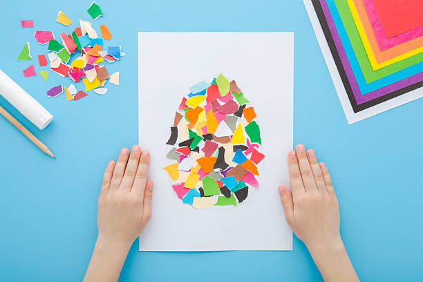 istock Little child hands showing glued easter egg shape from colorful torn application paper pieces on white paper. Light blue table background. Pastel color. Closeup. Point of view shot. Top down view. 2214181628