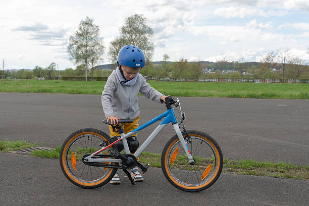 Young child in helmet balances bicycle, learning to ride on a paved path in a park. Bright day with clouds and trees in the background, focusing on effort and concentration.