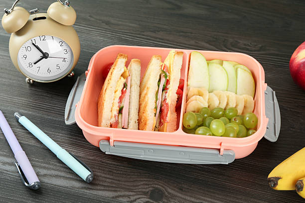 istock Lunch box with snacks, alarm clock and pens on wooden table 2214185817