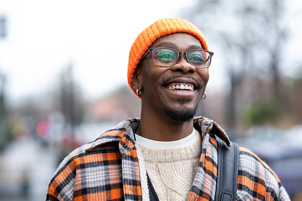 A close up Portrait of a stylish young man in a street, smiling warmly as he looks into the camera. He wears an orange and blue checkered jacket with a matching orange beanie, he has just finished thrifting at the charity shop, the image captures the fun and individuality of sustainable fashion.Videos are available similar to this scenario.