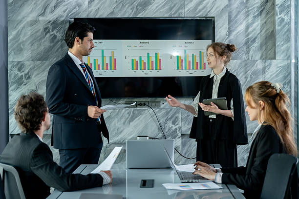 istock Multiracial business team having formal discussion in conference room while young businesswoman holding tablet presenting marketing data on screen with bar charts to group of colleagues 2214189505