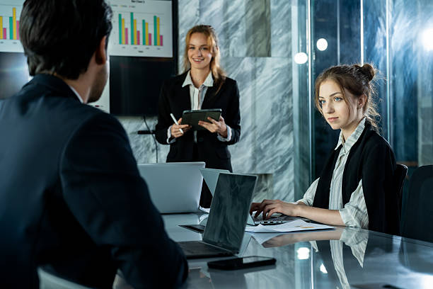 istock Young female business analyst using laptop working on report while other team members attending business presentation creating professional collaboration scene in corporate workspace 2214189507