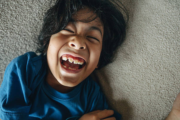 A young boy lying on a sofa in a cozy living room, laughing joyfully as a hand tickles him. He looks directly at the camera with a happy expression, capturing a playful, cheerful, and heartwarming family moment at home.