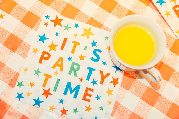 Kids party table with text It's party time on napkin and a vintage teacup with juice. Retro real-life candid image of children birthday party. Colorful patterns, stars and red checked tablecloth.