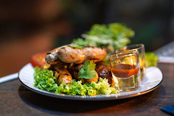 A plate of perfectly grilled sausages served on a bed of fresh lettuce, accompanied by roasted tomatoes, sauteed vegetables, and a side of mustard and chili sauce for dipping.