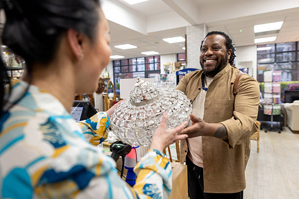 A medium over-the-shoulder shot of a man purchasing a glass light shade from a charity shop, located in Newcastle Upon Tyne. He is handing over the item to a female worker while smiling and wearing casual clothing. Videos are available similar to this scenario.