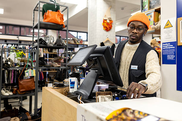 A wide shot of a man volunteering at a charity shop in Newcastle Upon Tyne. He is standing behind the cashier's desk while he tidies and organises everything around him. He is wearing casual clothing and an orange beanie.\n\nVideos are available similar to this scenario.