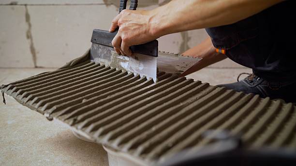 istock Laying tiles in the bathroom. Advertisement for tile adhesive. The spatula makes a comb mark on the tile. The craftsmen apply the adhesive solution to the tile with their hands, holding it in their hand. 2214233402