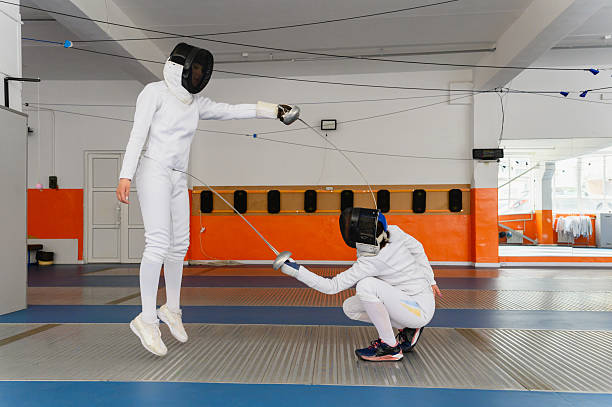 Female fencers in dynamic moment making simultaneous epee hit, one airborne, one crouching, side view, indoor hall with orange and white walls.