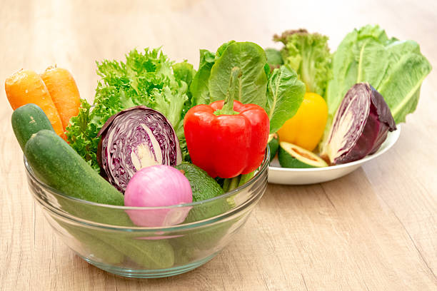 A colorful assortment of raw organic vegetables displayed on a light wood surface. The collection features crisp green lettuce, bright orange carrots, deep purple cabbage, a red bell pepper, a pink onion, and sliced avocado, highlighting healthy eating and fresh ingredients for a meal or snack. The scene is brightly lit, emphasizing the natural beauty and freshness of the produce.