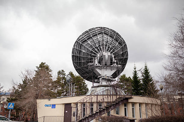 A large satellite dish stands on a platform surrounded by trees in an urban area, under a cloudy sky during the late afternoon hours.