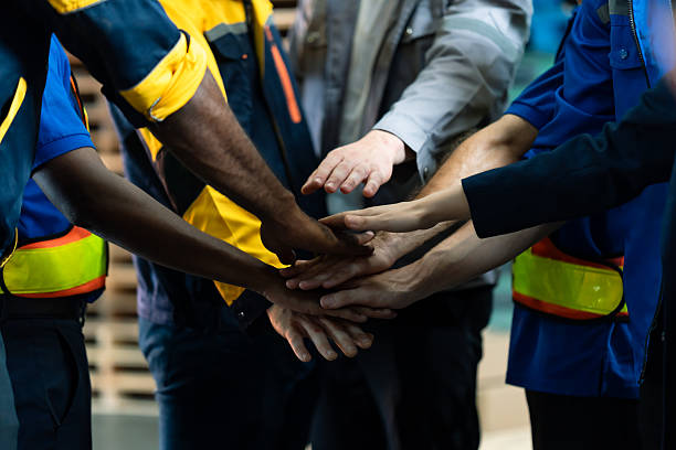 istock Group of logistics employees placing hands together in teamwork gesture, multiethnic group wearing safety uniforms in warehouse environment, industrial workplace unity and collaboration scene 2214248774
