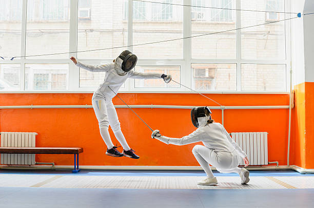 Female fencer airborne mid-jump as opponent crouches with extended epee, dynamic side view, bright indoor fencing hall with large window and orange walls.