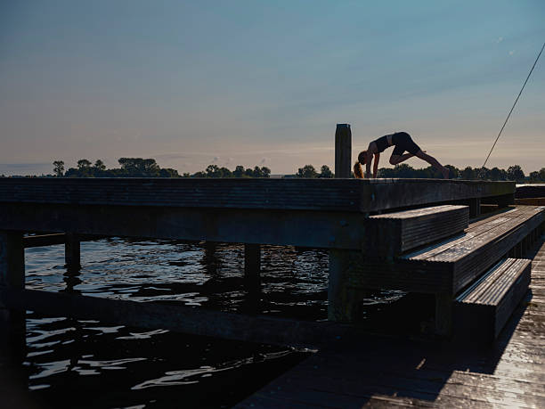 Dramatic silhouette of a person practicing yoga position on a wooden dock by a calm lake. The minimalist scene captures the tranquility of outdoor exercise at dusk or dawn, with the wooden platform extending over water and trees visible along the distant shoreline.