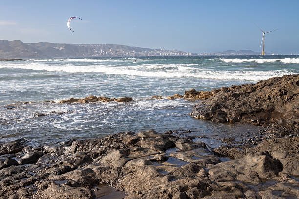 Rocky coast with boulders on a sandy beach of Atlantic ocean. Mountain and a wind turbine in the background. Waves with white foam. Blue sky. Telde, Las Palmas, Gran Canaria, Canary Islands, Spain.