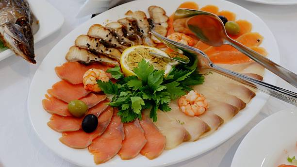 Catering service presenting a delicious fish platter with various sliced fish, shrimp, lemon, parsley, and olives arranged beautifully on a white plate with serving tongs