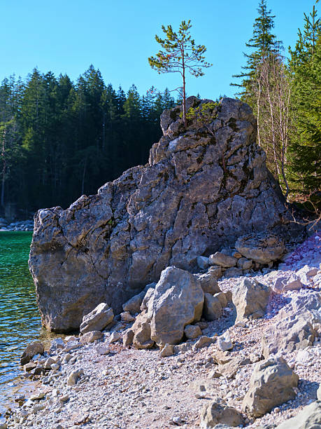 A unique rock formation stands by a calm turquoise lake under a clear blue sky. Lush forests surround the area, highlighting the beauty of nature in this serene location.