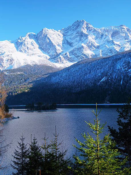 Snow-covered peaks tower above a tranquil lake, capturing the beauty of winter. Evergreen trees frame the scene, enhancing the calm atmosphere on a bright day.
