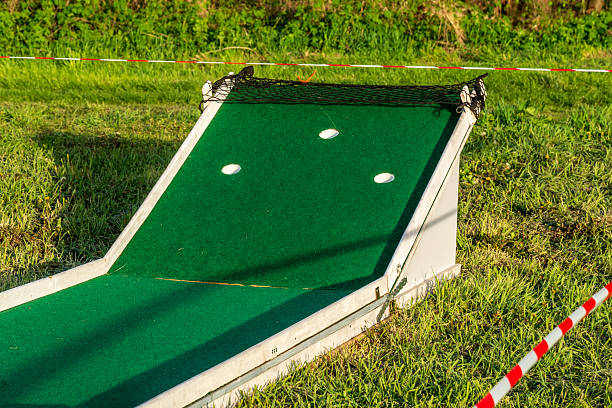 Close-up of a mini golf obstacle with green turf, three holes and netting, set on a grassy area with safety tape.