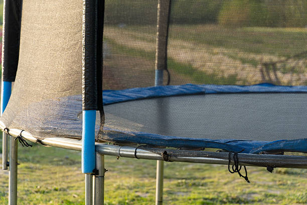 A close view of a trampoline with safety netting and blue padding, set outdoors on a grassy lawn. Recreation, activity, play.