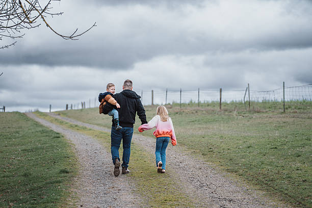 Rear view of a farmer carrying his son on his hip and holding hands with his elementary age daughter as they walk together down a gravel road on their farm in Oregon where they grow root vegetables.