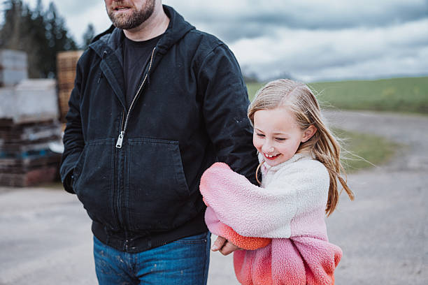 A smiling elementary age girl affectionately holds her dad's arm as they walk together outside at the agricultural farm their family owns in rural Oregon, USA.