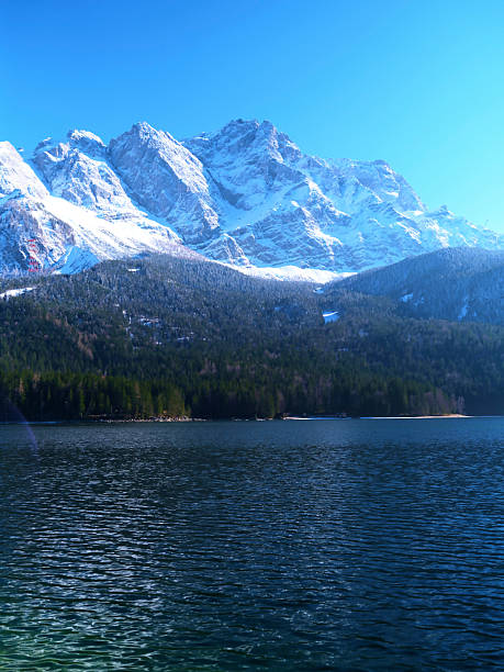Snow-capped peaks rise above a serene lake, surrounded by lush evergreen forests, showcasing nature's beauty on a clear, sunny day.