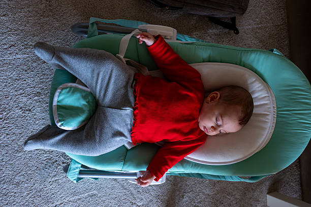 A baby wearing a red shirt and gray pants peacefully sleeps in a green baby rocker inside a cozy room.