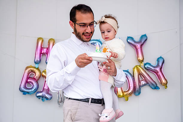 Father joyfully holds his baby and a number one candle cake during their first birthday celebration, standing in front of a vibrant balloon display spelling 'Happy Birthday.' The atmosphere is cheerful and celebratory.