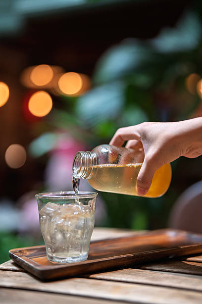 A hand pouring a chilled beverage from a glass bottle into an ice-filled glass, set on a wooden tray with a bokeh garden background.