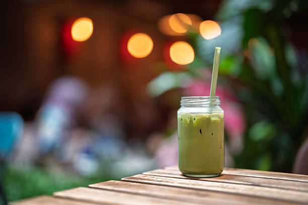 A refreshing iced matcha latte served in a glass jar with a straw, placed on a rustic wooden table in a cozy outdoor setting with warm bokeh lights in the background.